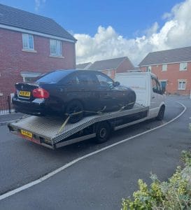 Black saloon car strapped onto a flatbed recovery truck in a housing estate