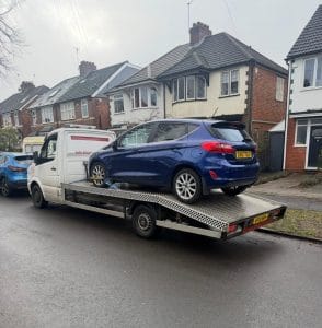 Blue hatchback loaded onto a flatbed recovery truck on a residential street