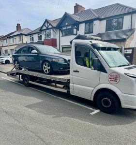 Dark saloon car secured on a flatbed recovery truck outside residential homes