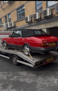 Red convertible car secured on a flatbed recovery truck outside a commercial building