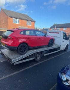 Red saloon car secured on a flatbed recovery truck outside homes