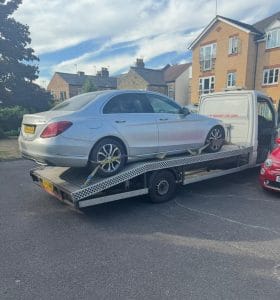 Silver saloon car loaded onto a tilted flatbed recovery truck in a residential area