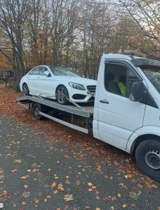 White saloon car secured on a flatbed recovery truck beside autumn trees