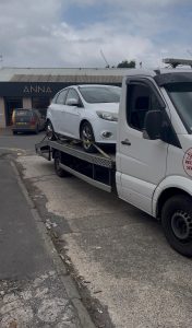 White van secured on a recovery truck on a roadside in an urban area