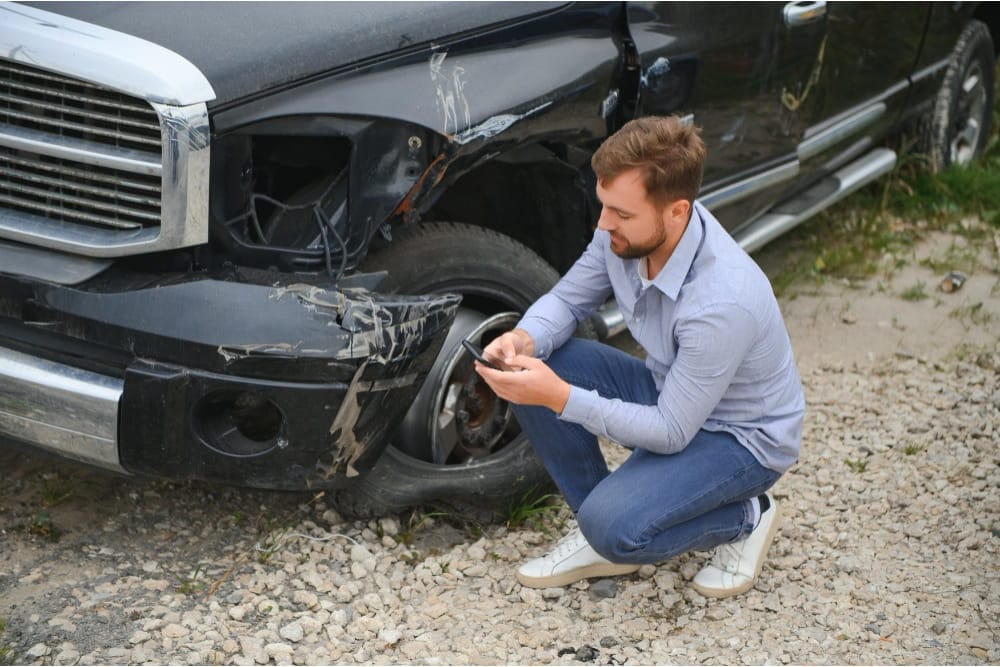 A man uses his phone for accident recovery next to a damaged black truck